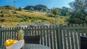 A dining area with a table and chairs, a wooden fence, and a view of a hill with rocks and trees.