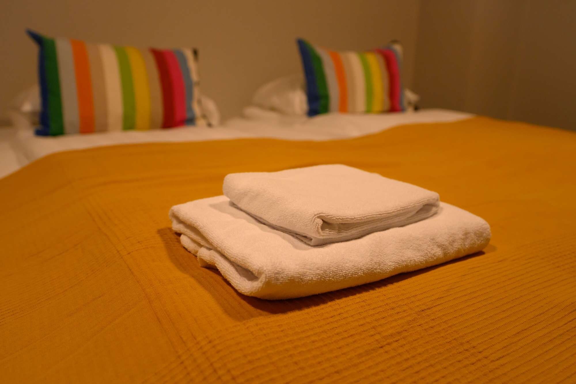 Ein Bett mit einem gefalteten weißen Handtuch, einem gestreiften Kissen und einer schlichten Wand im Hintergrund.