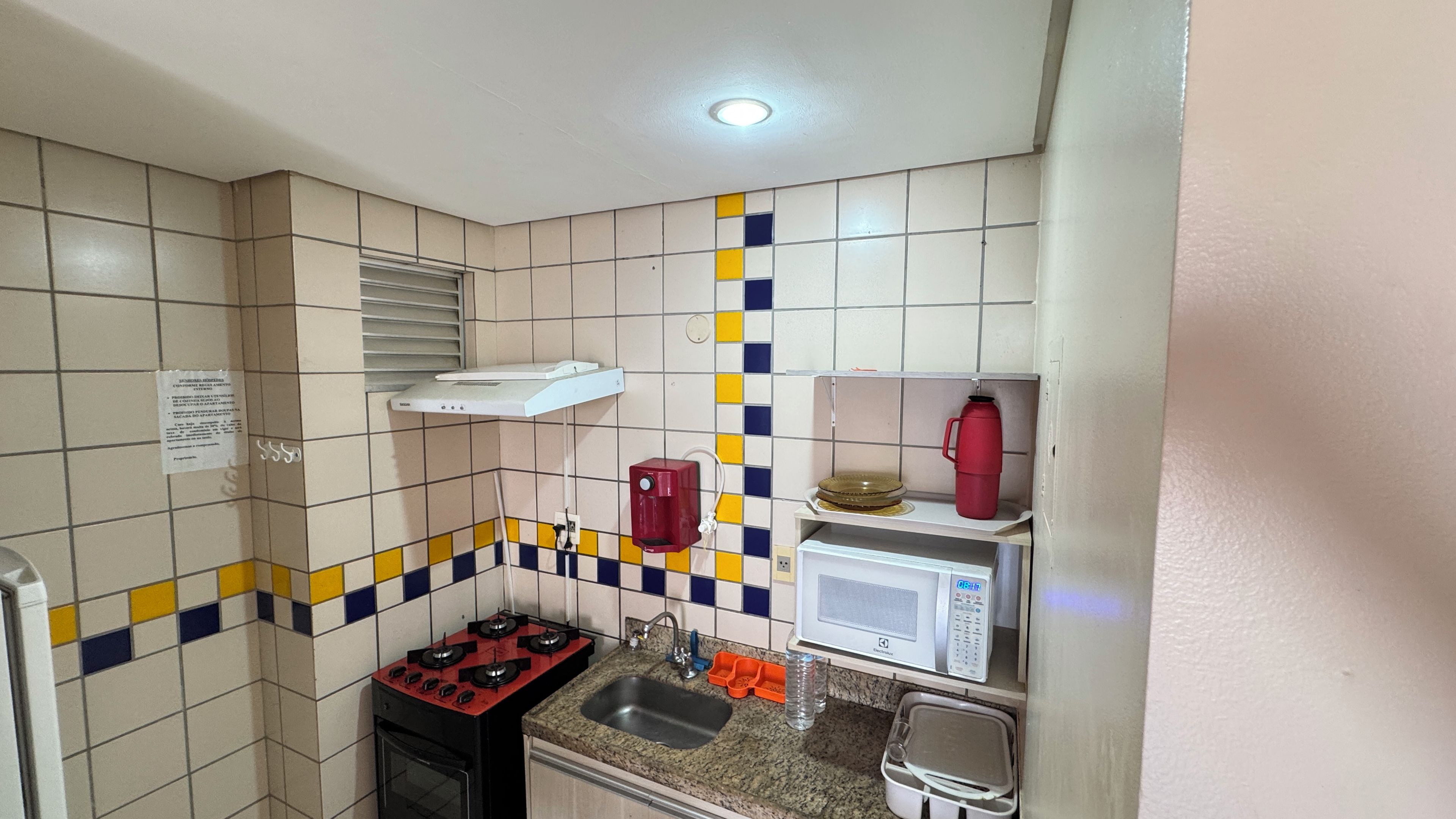 A compact kitchen with a gas stove, a microwave, a red water dispenser, and a tiled backsplash.