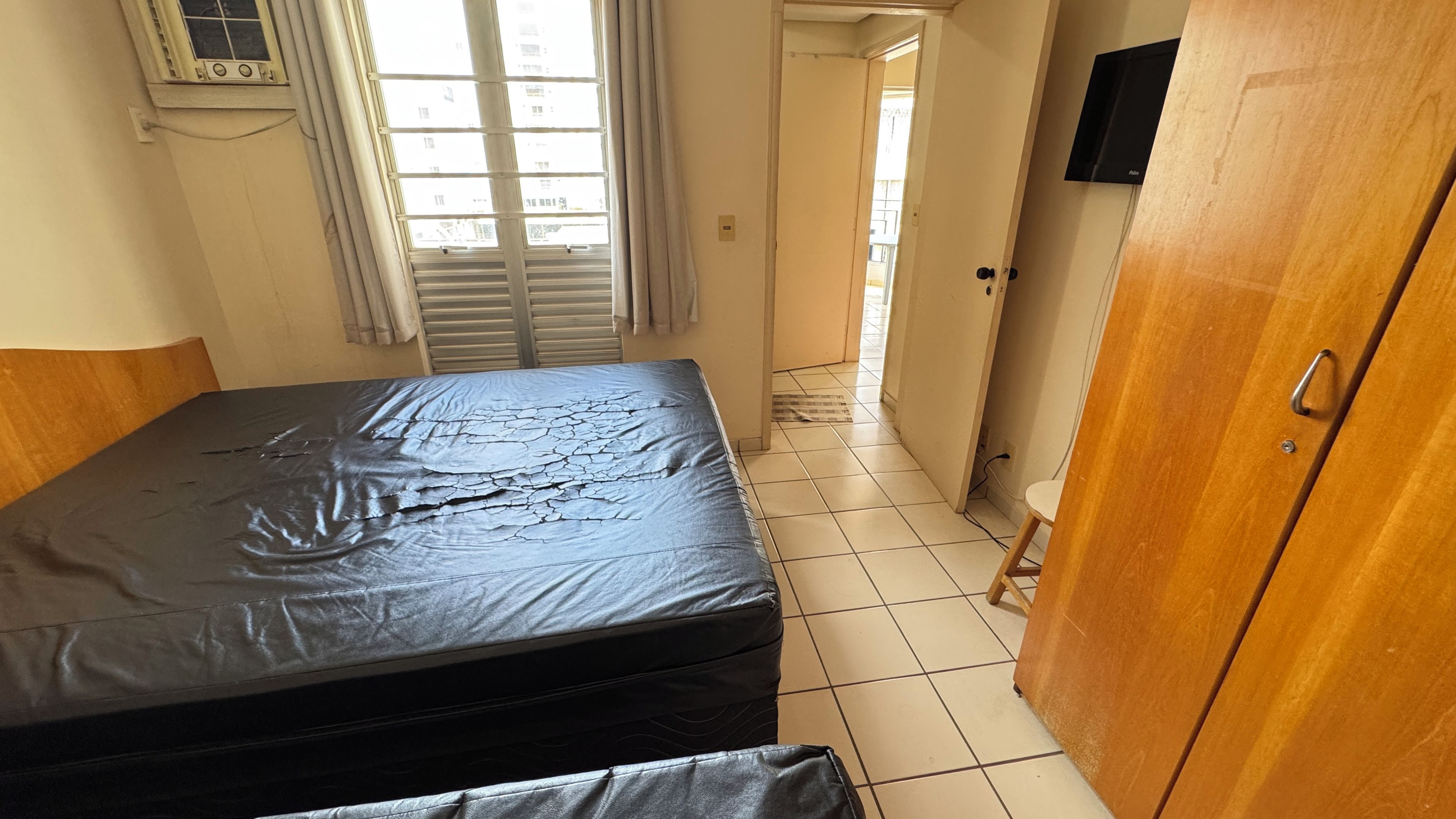 Une chambre avec un lit, une armoire en bois, un téléviseur fixé au mur et une fenêtre avec des rideaux.