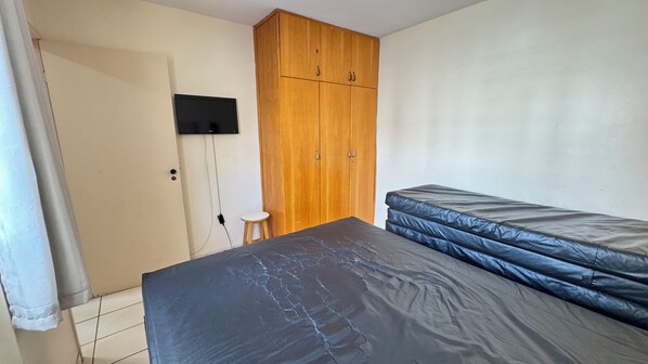 A bedroom with a bed, a wooden wardrobe, a small stool, and a television mounted on the wall. - CRCN - Eldorado Park (Caldas Novas)