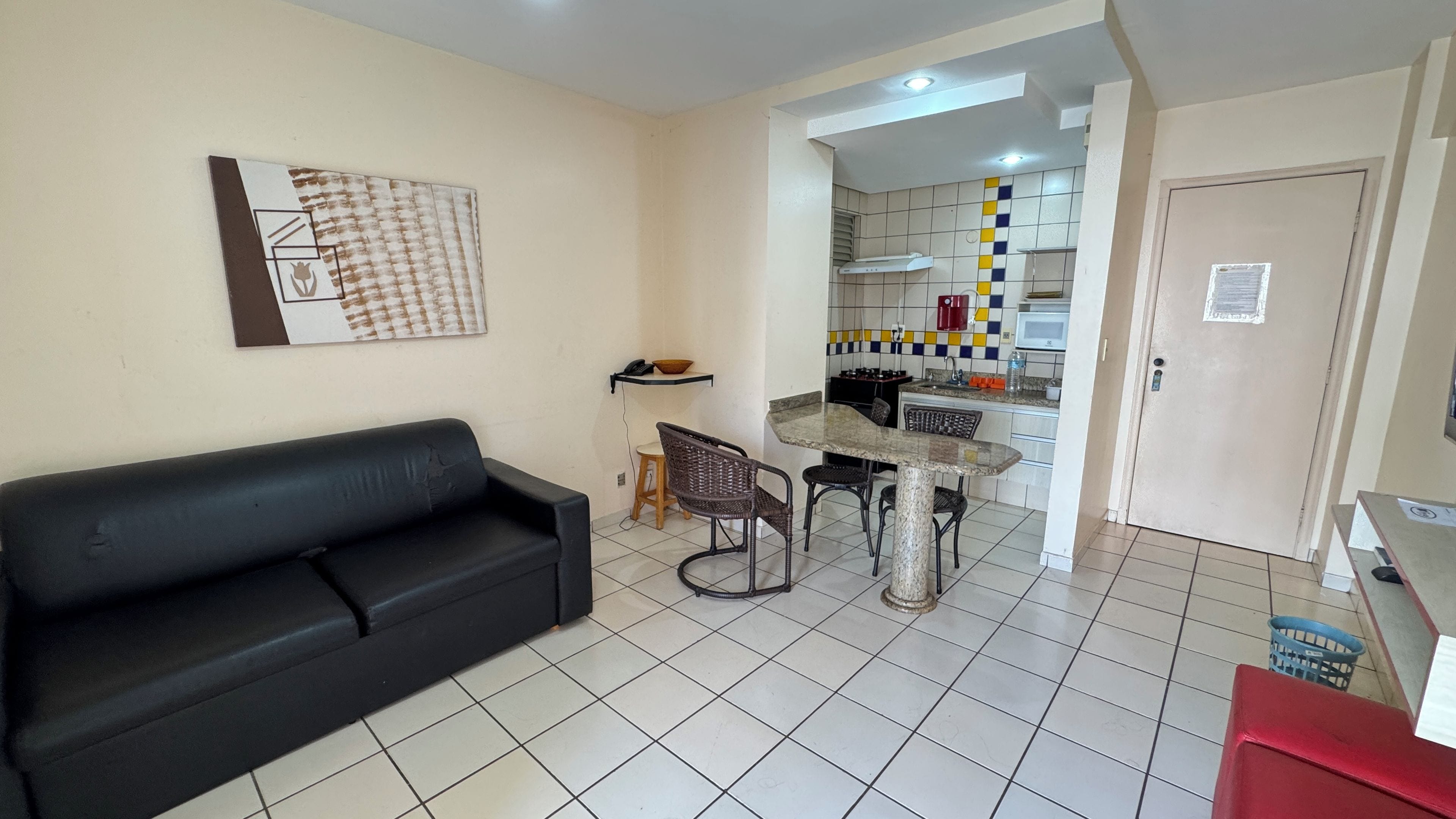 Uma sala de estar com um sofá preto, uma mesa de jantar com quatro cadeiras e uma área de cozinha com micro-ondas e fogão.