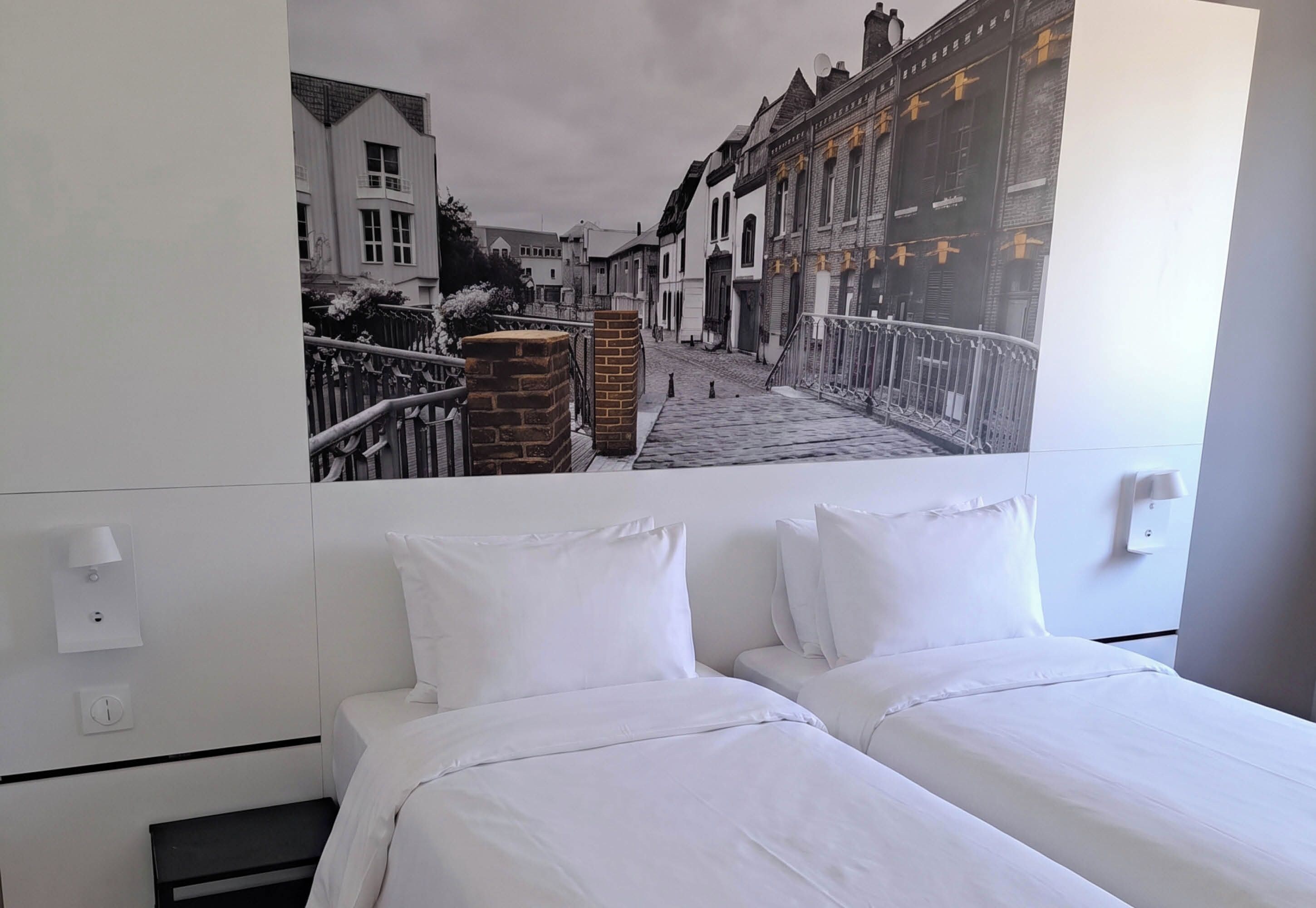 A hotel room with a bed, a wall-mounted lamp, and a large window.
