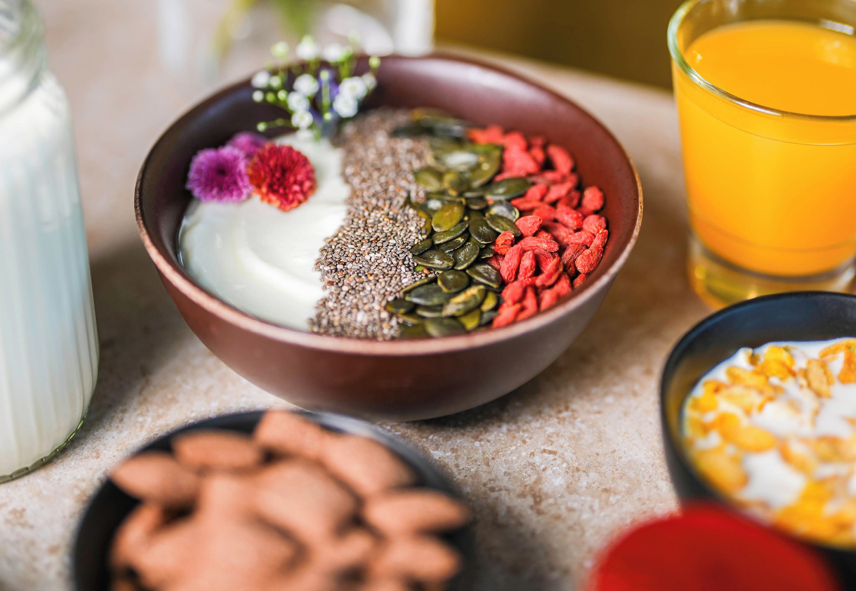 Un cuenco con semillas y flores secas, un vaso de jugo de naranja y una botella de leche sobre una encimera.