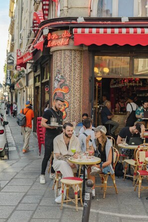 An outdoor cafe with wicker chairs and tables, a street view with pedestrians, and a building with a red awning and a neon sign. - Elegant Saint-Germain Apartment with Refined Parisian Interiors (Paris)