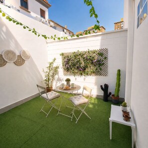 Une terrasse avec un revêtement de gazon synthétique, deux chaises, une petite table, des assiettes décoratives, un cactus et un mur en treillis sur lequel grimpent des plantes.