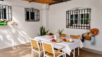 A dining area with a table set for breakfast, surrounded by chairs, with a white tablecloth, and a view of the outside through barred windows.