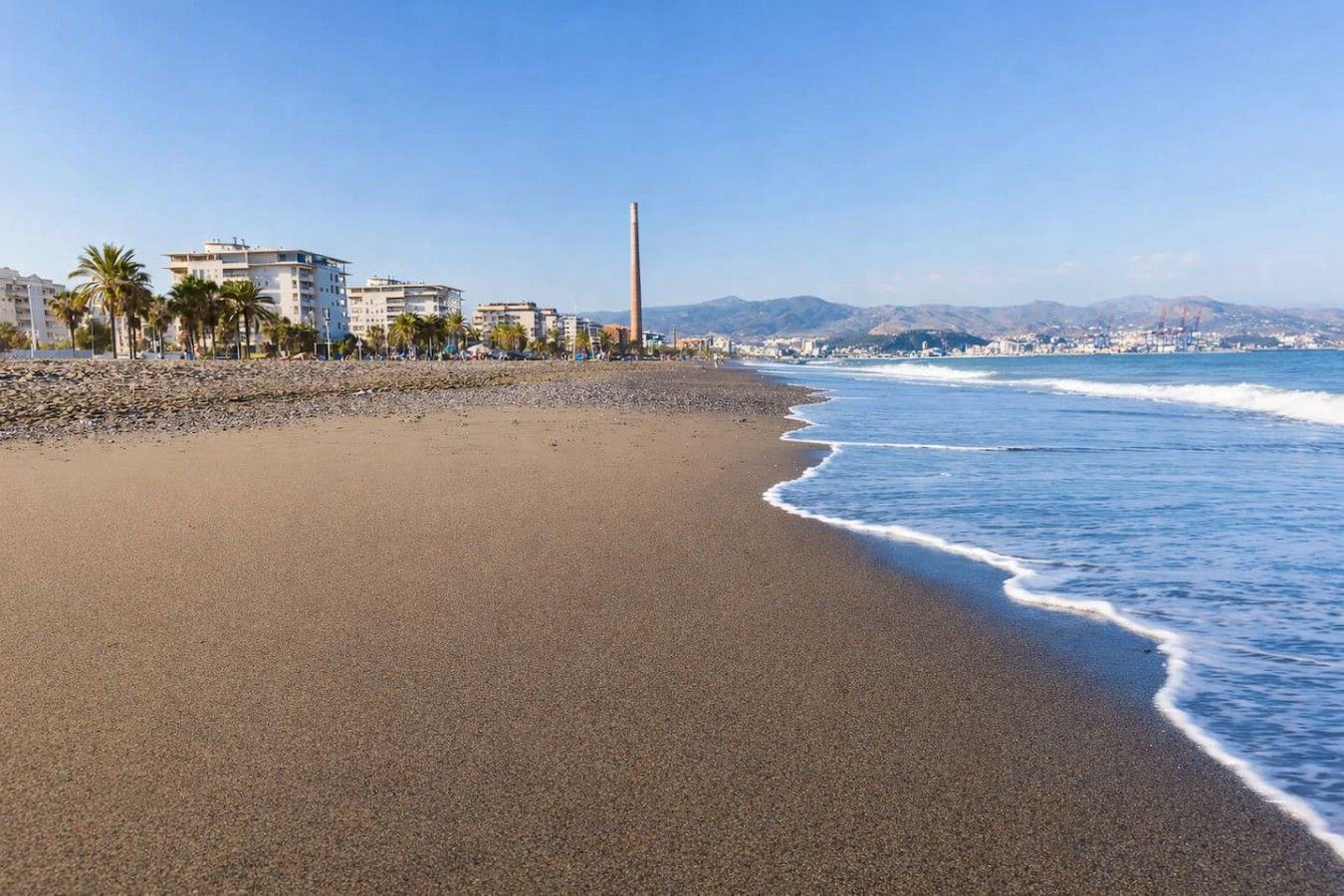 A beach with a clear sky, a sandy shore, and buildings in the background.