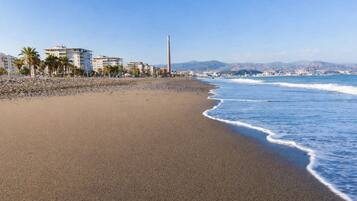 A beach with a clear sky, a sandy shore, and buildings in the background.