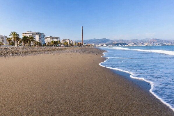 A beach with a clear sky, a sandy shore, and buildings in the background. - 2 bedrooms | Large patio | 2-meter walk to the beach | Huelin (Málaga)