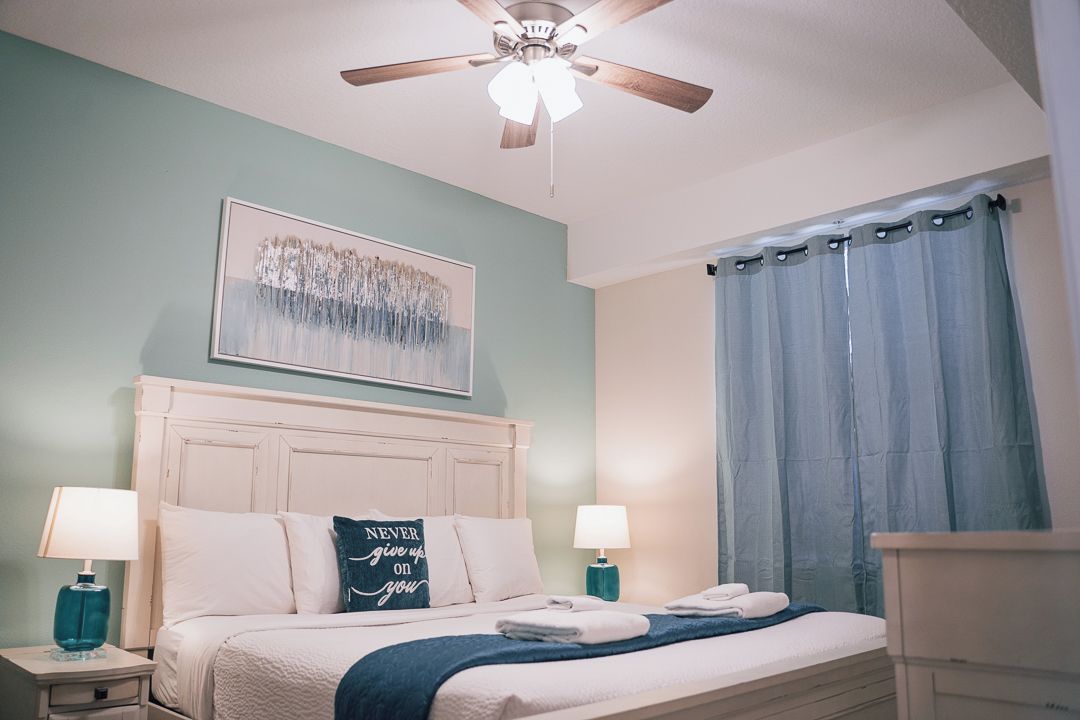 Un dormitorio con cama, mesitas de noche, ventilador de techo, un cuadro y una ventana con cortinas.