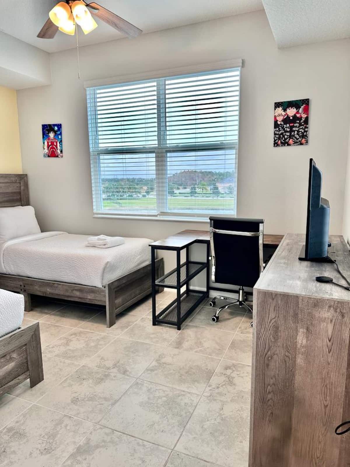 A bedroom with a bed, a desk, and a chair. There is a window with blinds, a ceiling fan, and artwork on the walls.