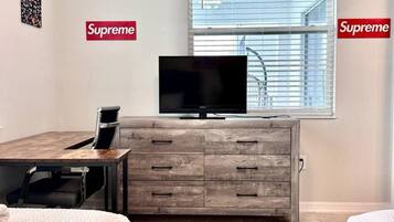 A bedroom with a bed, a TV on a wooden cabinet, and a window with blinds.