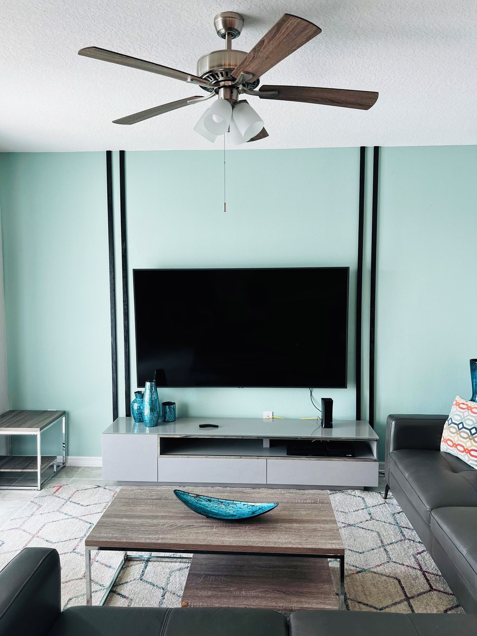 A living room with a flat-screen TV, a ceiling fan, a coffee table, a sofa, and a side table.