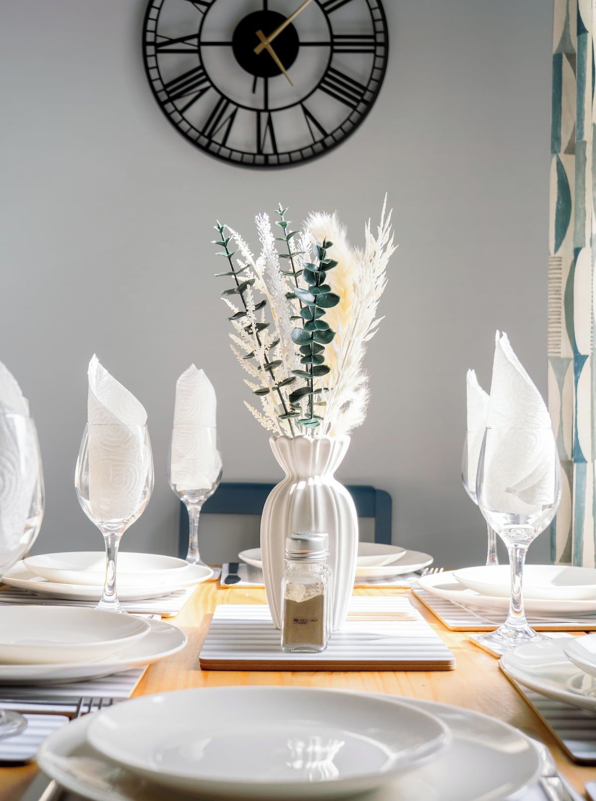 Ett matbord dukat med vita tallrikar, glas och en vas med torkade blommor.