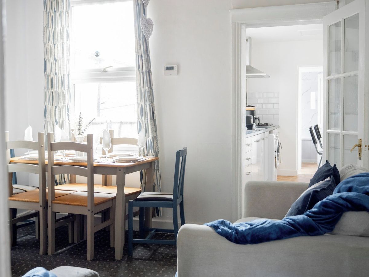 A room with a dining table set for two, a sofa, and a kitchen area visible through an open door.