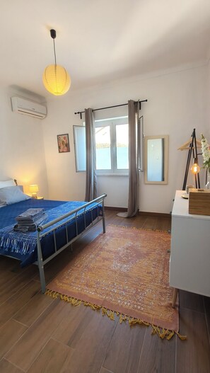 A bedroom with a bed, a window with curtains, an air conditioning unit, a bedside lamp, and a bedside table with a lamp. - 2-bedroom house, office with sofa-bed, air con, wifi, garden shower, terrace (Cano)