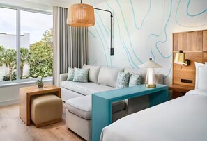 Ein modernes Hotelzimmer mit einer Couch, einem kleinen Tisch, einem Bett und einem Nachttisch. Es gibt ein Fenster mit Blick auf Grünflächen.