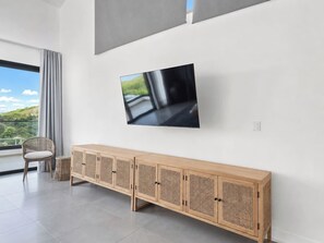 A modern living room with a flat-screen TV mounted on the wall, a wooden console table with multiple compartments, a wicker chair, and a large window offering a view of greenery. - Villa La Mar | Golf & Ocean Views, Private Pool (Punta Cana)