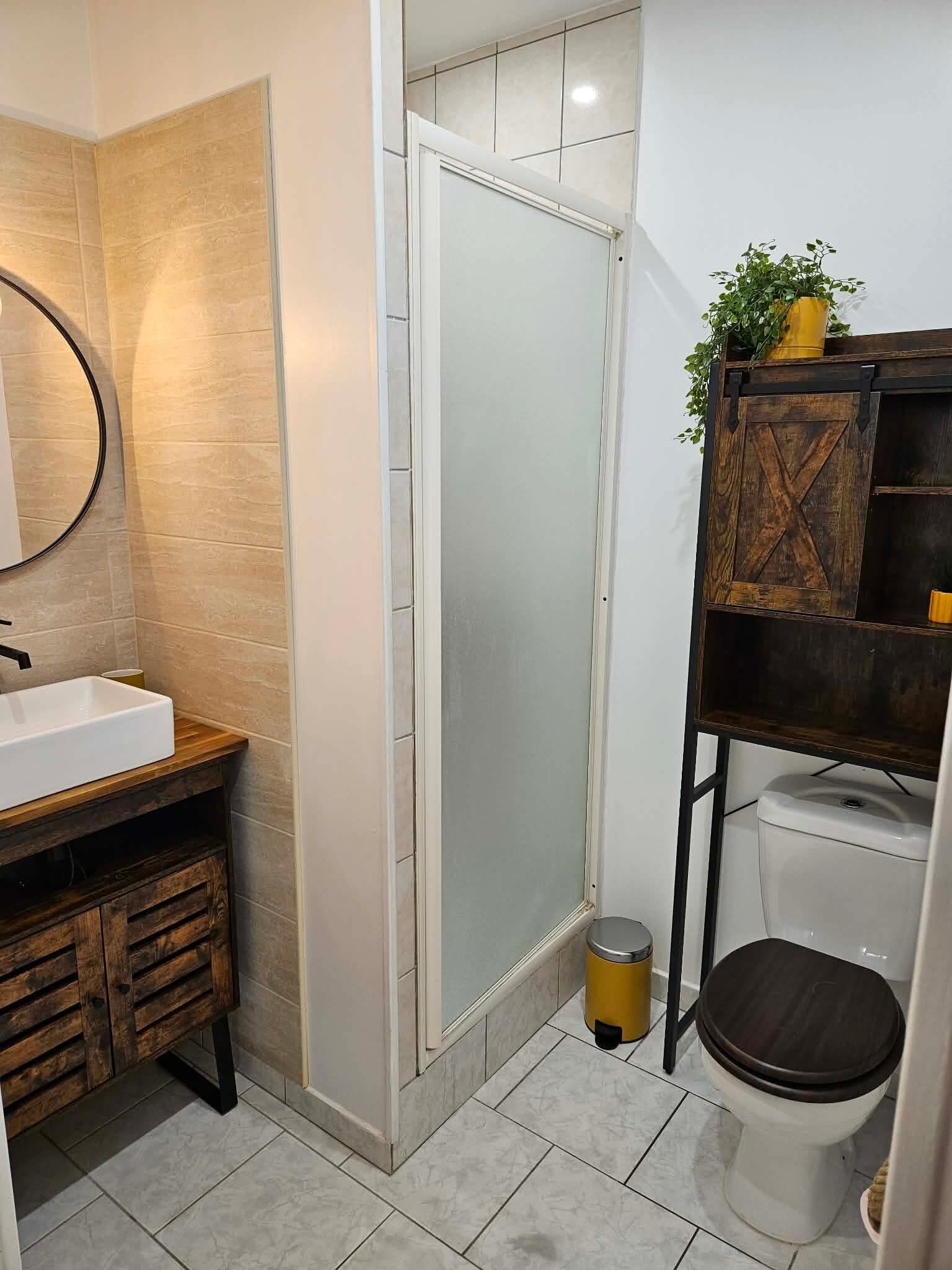 A bathroom with a wooden vanity, a white sink, a mirror, a frosted glass shower door, a toilet, and a small trash bin.