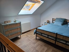 A bedroom with a skylight, a bed with blue bedding, a dresser with wicker baskets, and a small bedside table with a lamp.