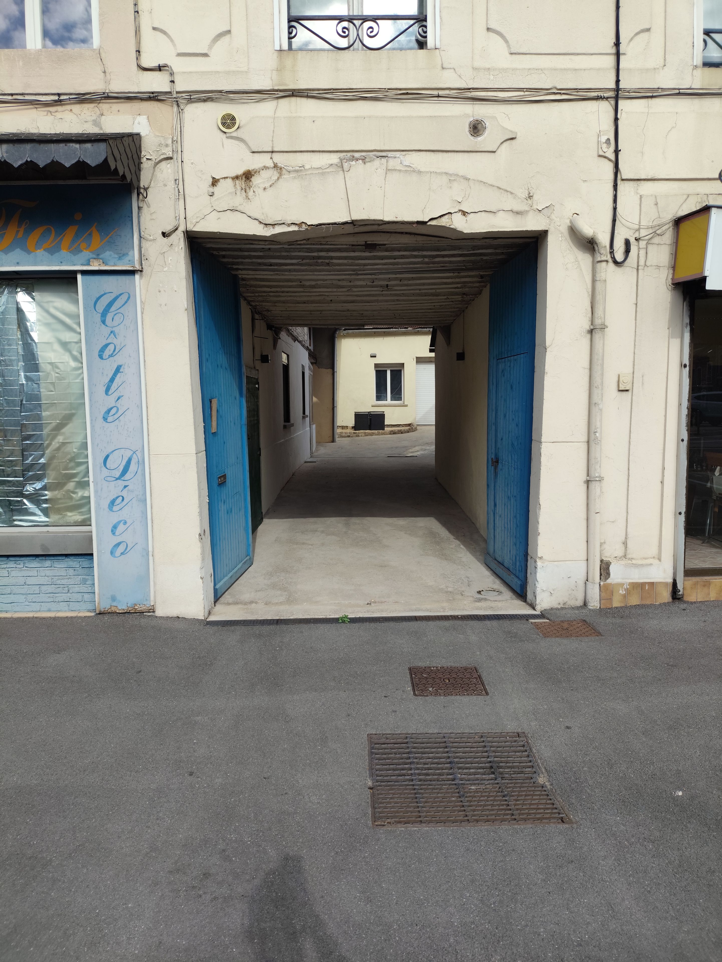 Un couloir avec une porte bleue à gauche et une porte blanche à droite, menant à un bâtiment doté d’une fenêtre et d’une porte.
