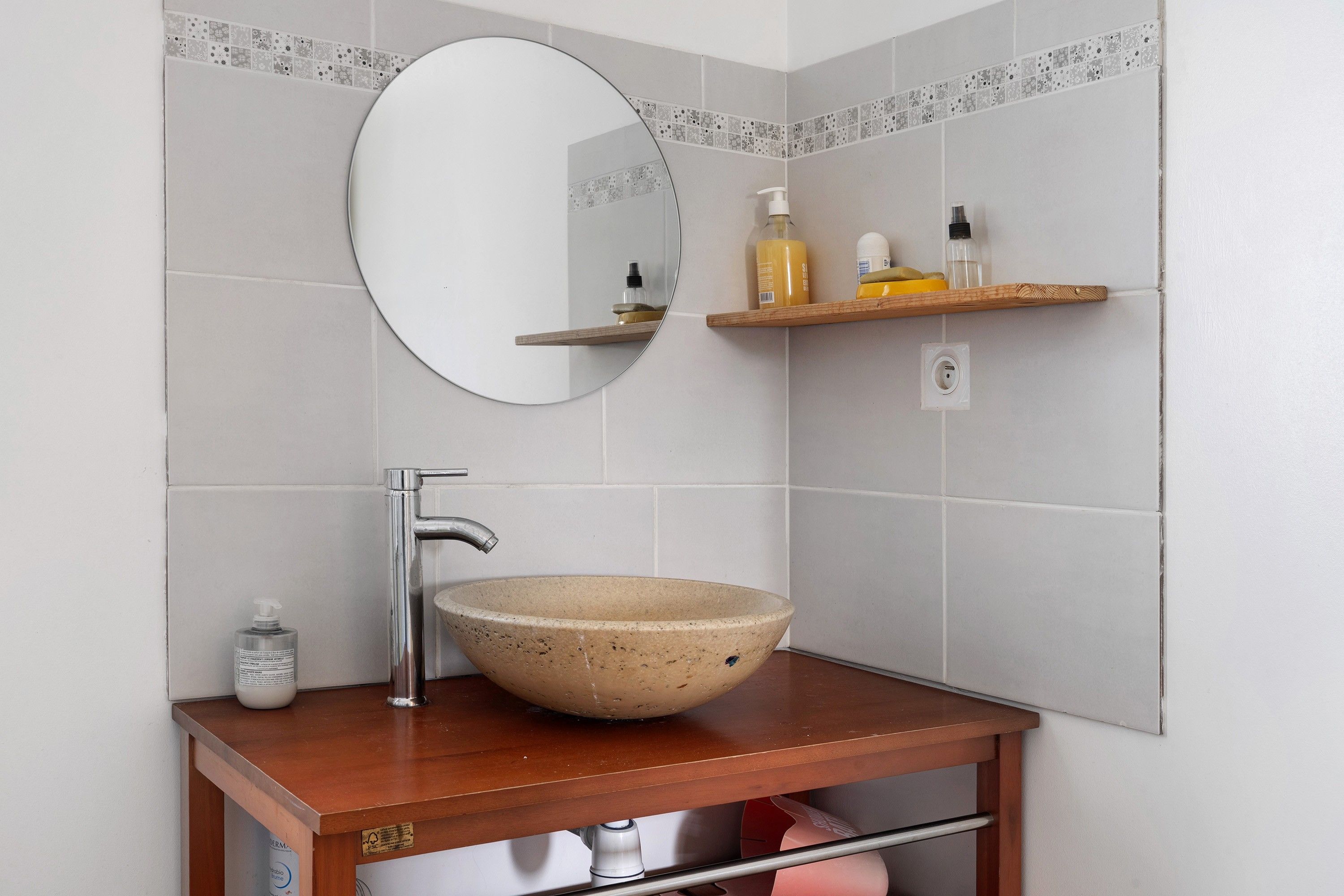 Een badkamer met een ronde spiegel, een beige keramieken wastafel, een houten plank met toiletartikelen en een handdoekhouder.