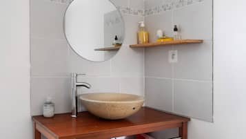 Een badkamer met een ronde spiegel, een beige keramieken wastafel, een houten plank met toiletartikelen en een handdoekhouder.
