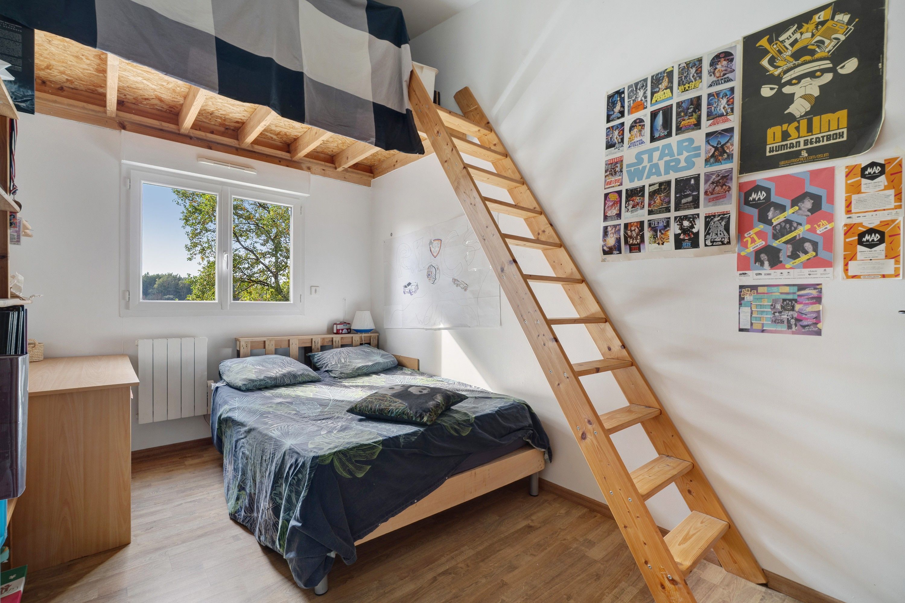 Une chambre avec un lit superposé, une échelle en bois, une fenêtre et un mur décoré d’affiches.