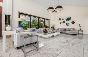 A spacious living room with a large window, a sofa set, a coffee table, and decorative items. - Catalina 6: Luxury retreat w/ pool, cinema, gym, golf carts & staff (Casa de Campo)