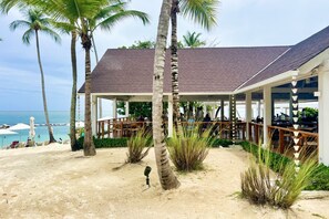 A beachside building with a covered patio, palm trees, and a view of the ocean. - Catalina 6: Luxury retreat w/ pool, cinema, gym, golf carts & staff (Casa de Campo)