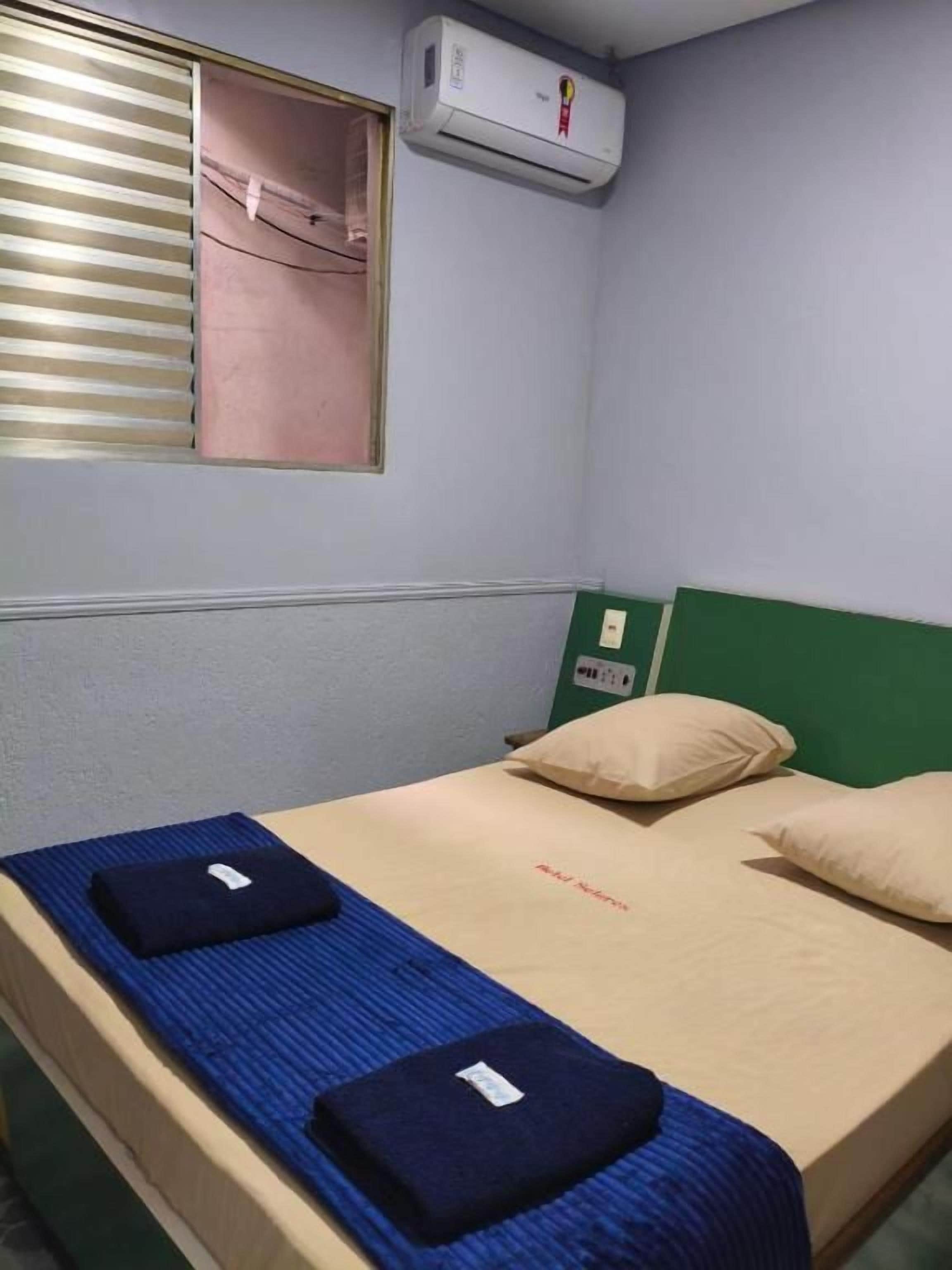 Uma cama de solteiro com cabeceira verde, janela com persianas e um aparelho de ar condicionado fixado na parede.