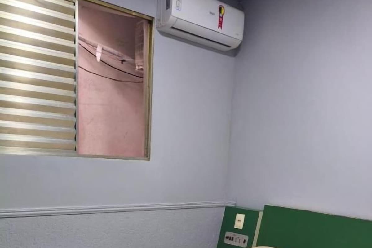 Una cama individual con cabecera verde, una ventana con persianas y un equipo de aire acondicionado instalado en la pared.