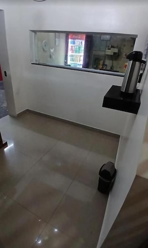 A room with a tiled floor, a black trash bin, a black shelf with a water dispenser, and a window with a vending machine. - Hotel solares  (São Paulo)