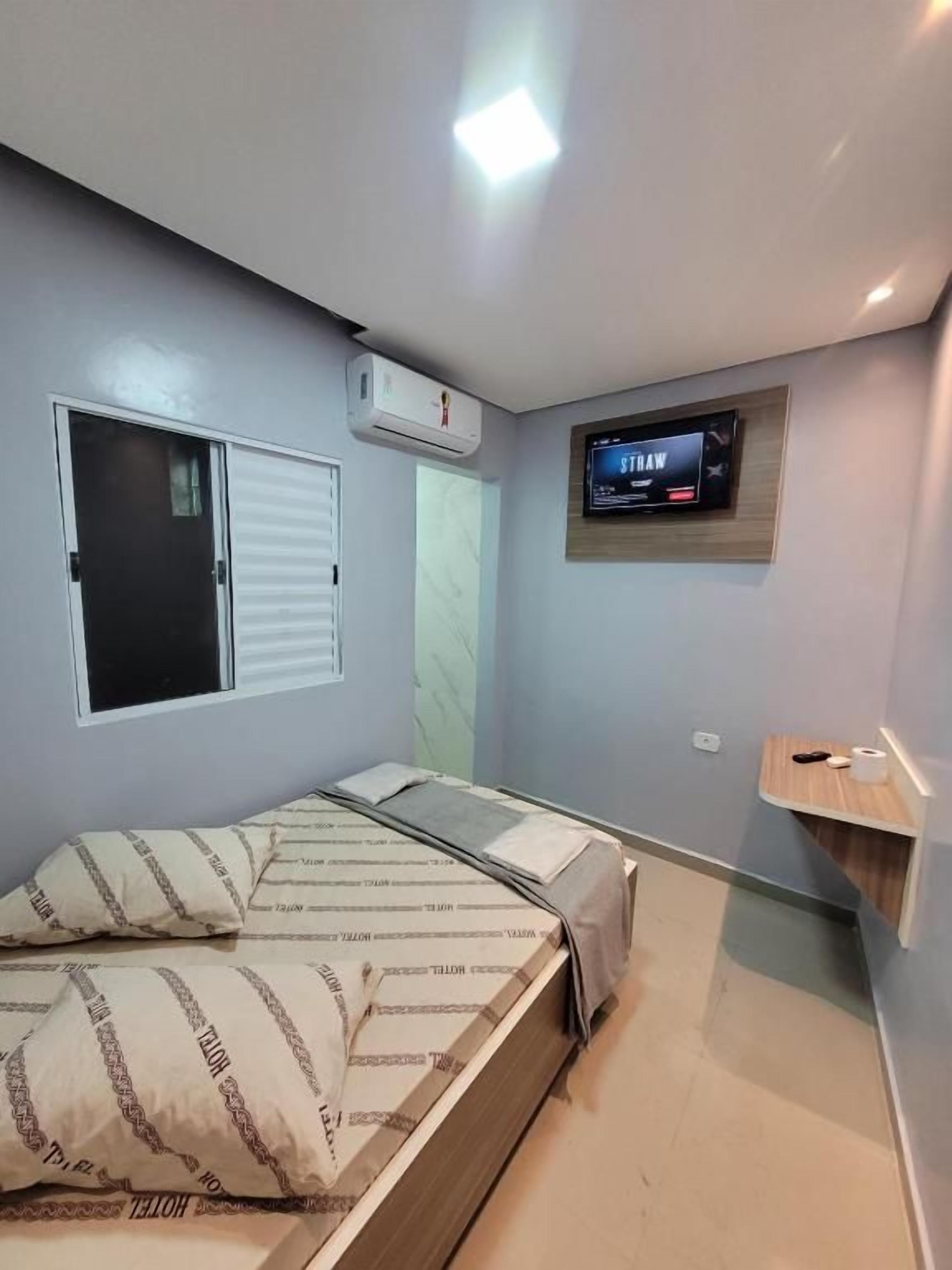 A bedroom with a bed, a TV mounted on the wall, and a window with blinds.
