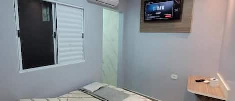 A bedroom with a bed, a TV mounted on the wall, and a window with blinds.