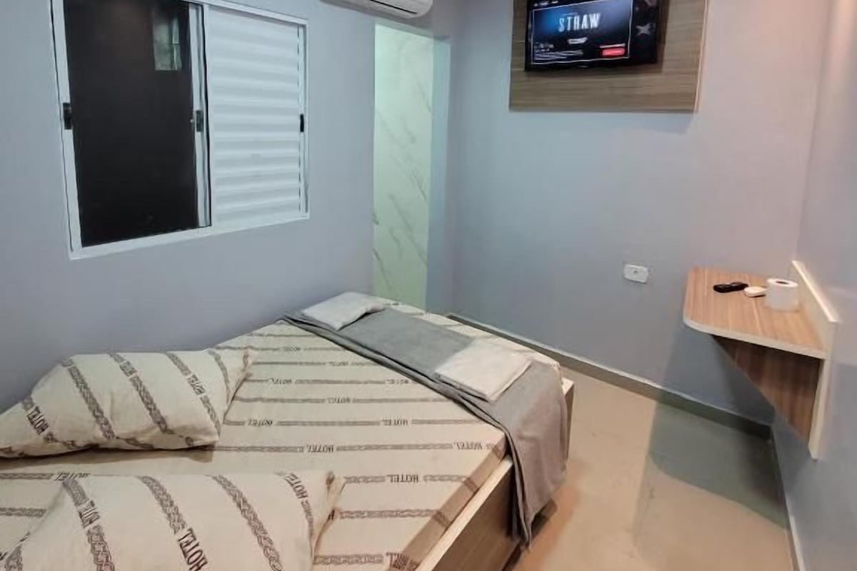 Un dormitorio con cama, televisor montado en la pared y ventana con persianas.