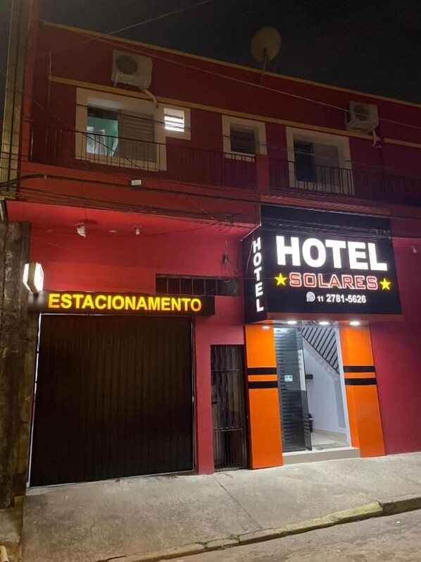 A hotel with a prominent sign reading 'HOTEL SOLARES' and a contact number. There is a closed door labeled 'ESTACIONAMIENTO' and a staircase leading to the entrance.