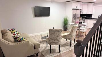 A living room with a sofa, two armchairs, a flat-screen TV, a dining area with a table and chairs, and a kitchen with white cabinets and appliances.