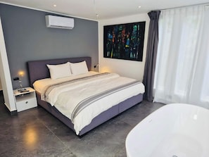Una habitación de hotel moderna con una cama grande, una mesita de noche, un aire acondicionado de pared, un ventanal con cortinas y una bañera.