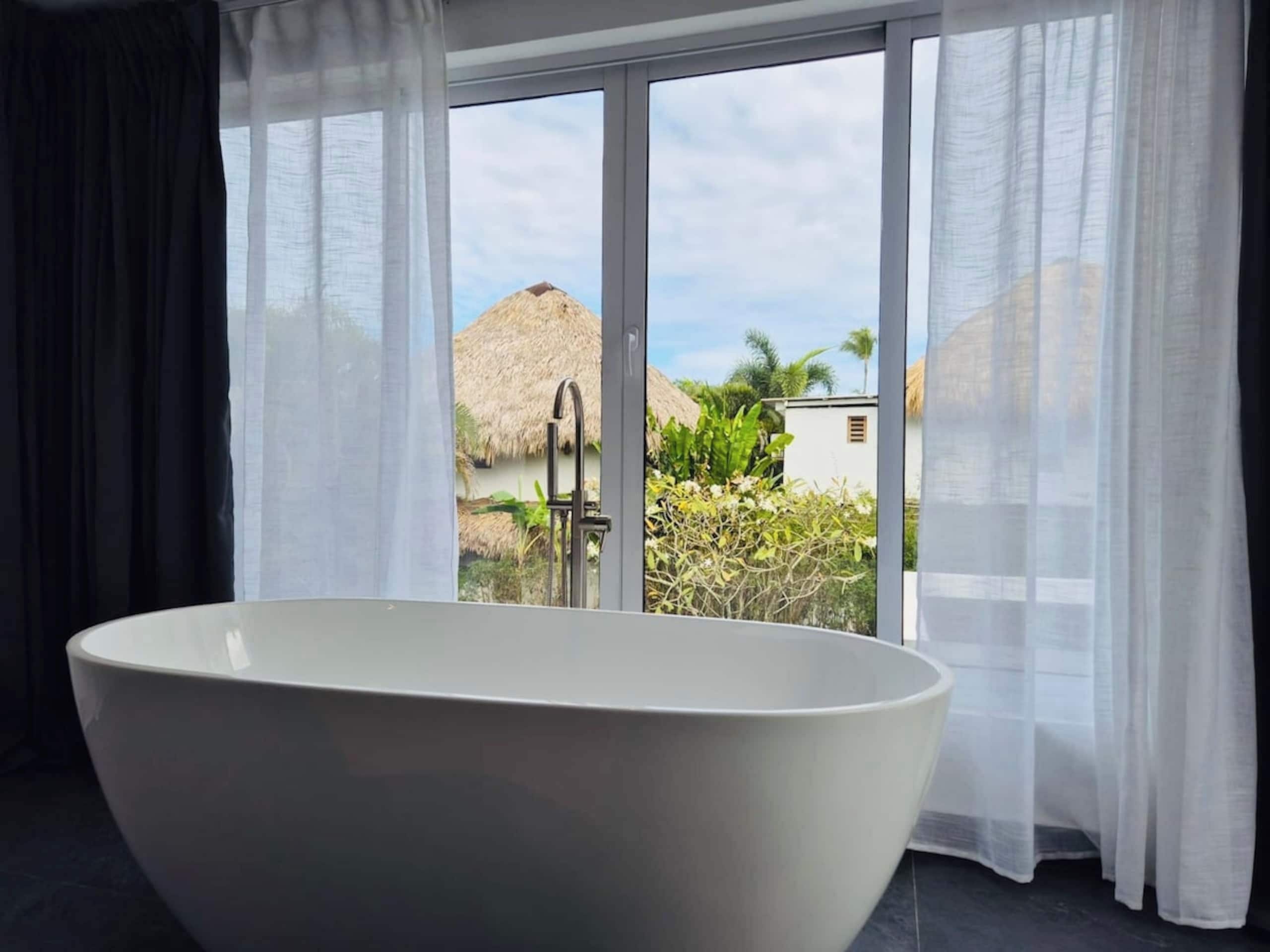 A modern bathroom with a freestanding bathtub, large windows with sheer curtains, and a view of a tropical landscape.