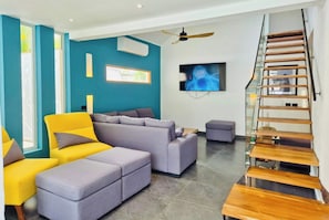 Una sala de estar moderna con un sofá seccional, un sillón amarillo y un reposapiés gris. Se ve una escalera de madera con barandas de vidrio a la derecha.