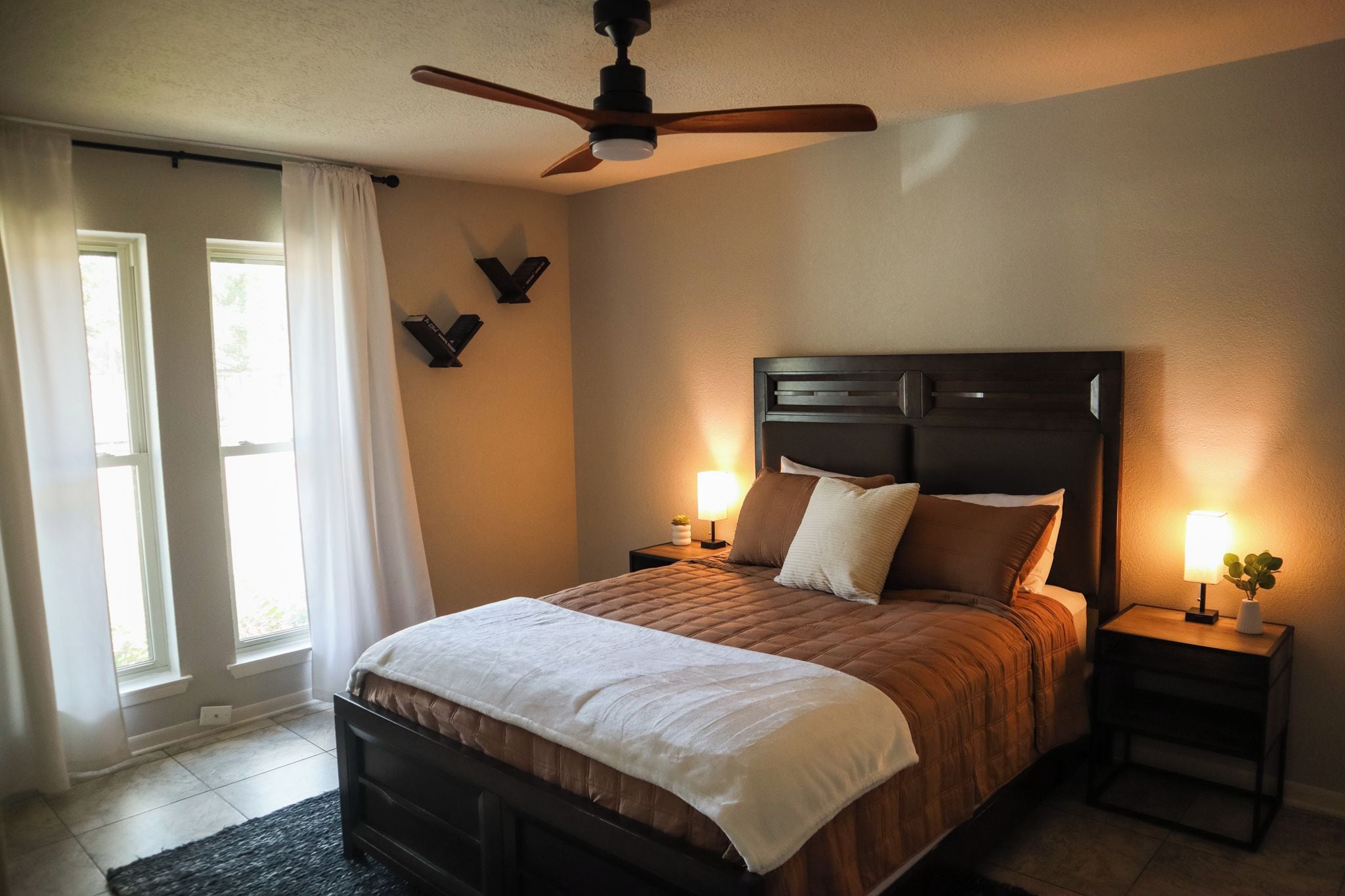 A bedroom with a bed, two bedside tables, a ceiling fan, and a window with curtains.
