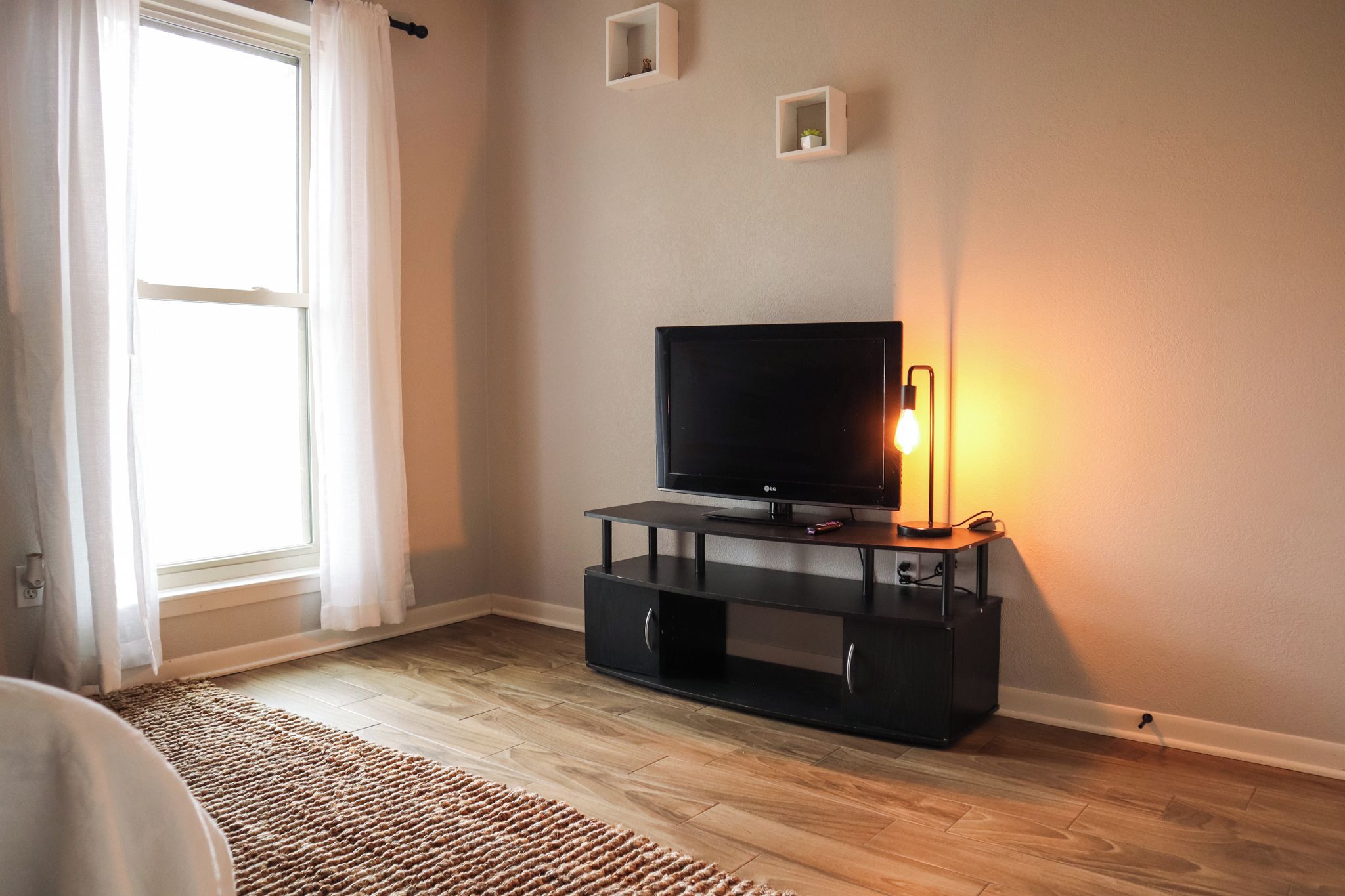 A living room with a flat-screen TV on a black stand, a small table with a lamp, and a window with curtains.