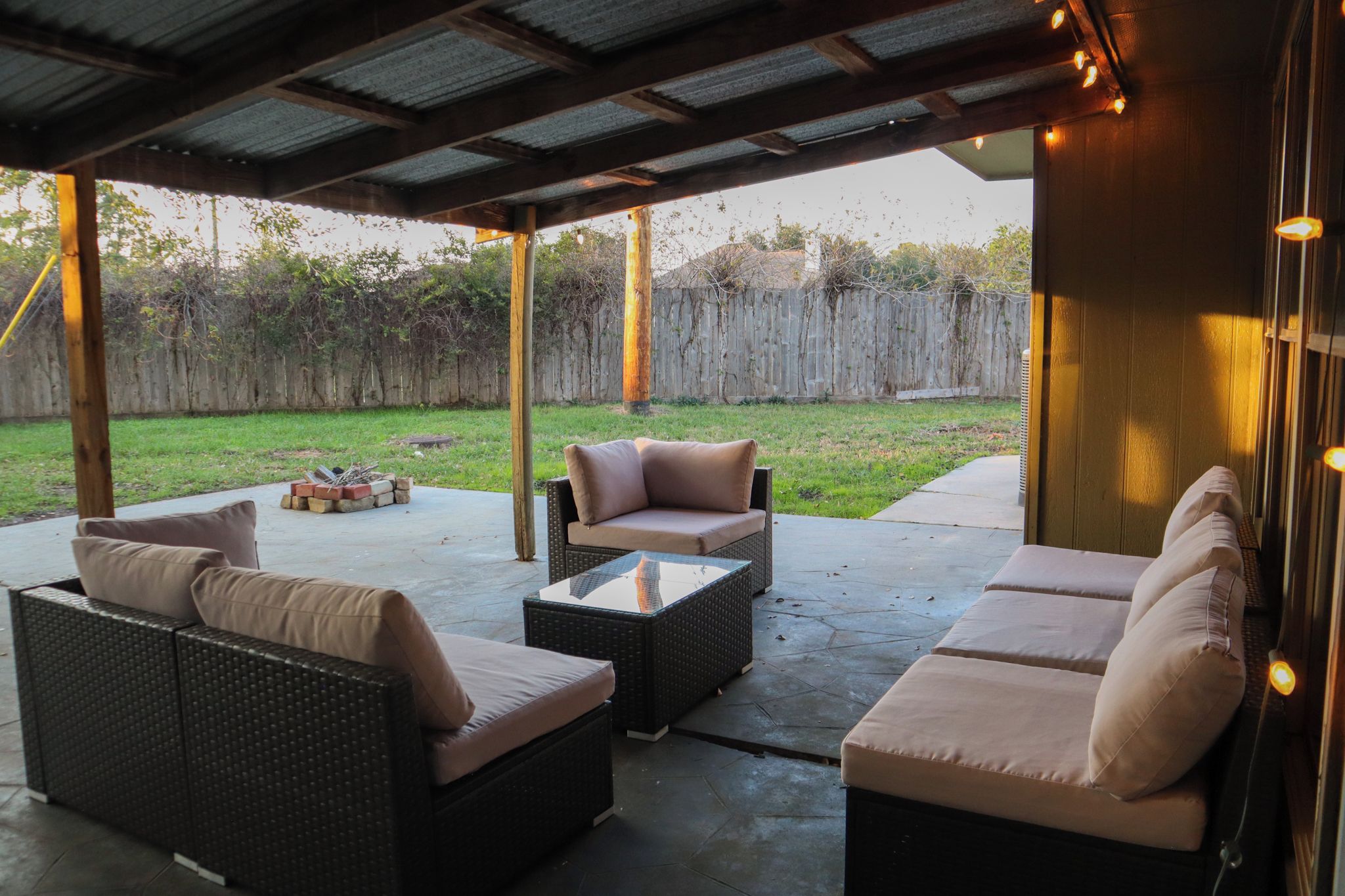 An outdoor patio with wicker furniture, a glass-top coffee table, and string lights.
