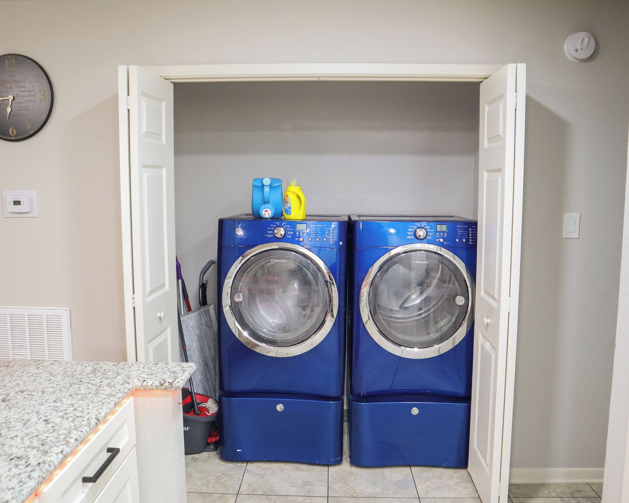 A laundry room with two blue front-loading washing machines and a small shelf with cleaning supplies.