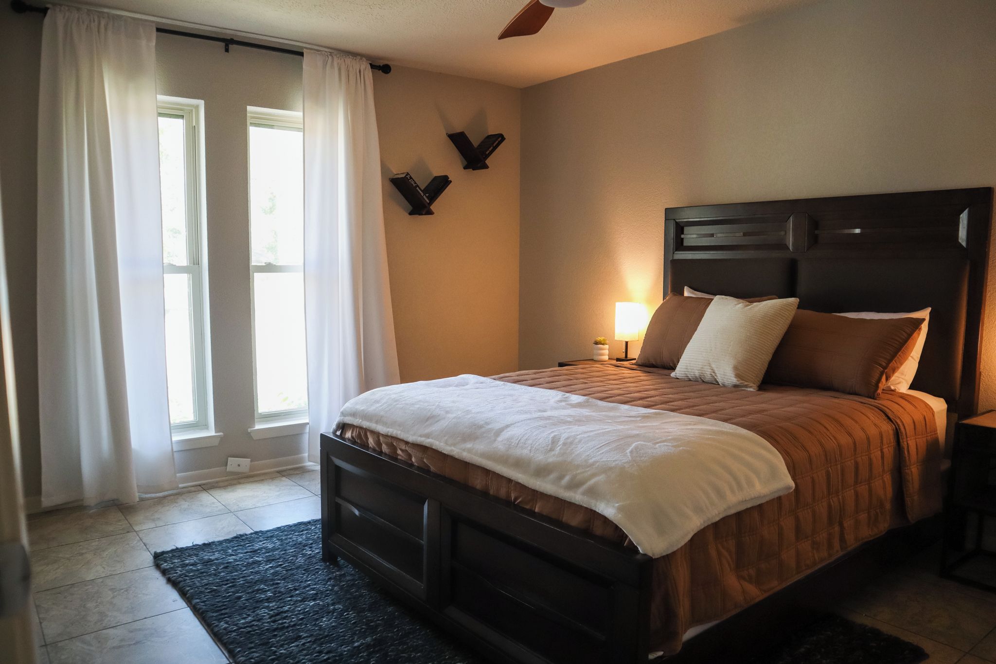Quarto com cama, mesa de cabeceira, abajur e janela com cortinas.