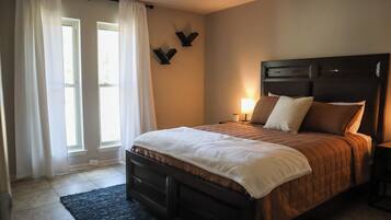 Quarto com cama, mesa de cabeceira, abajur e janela com cortinas.