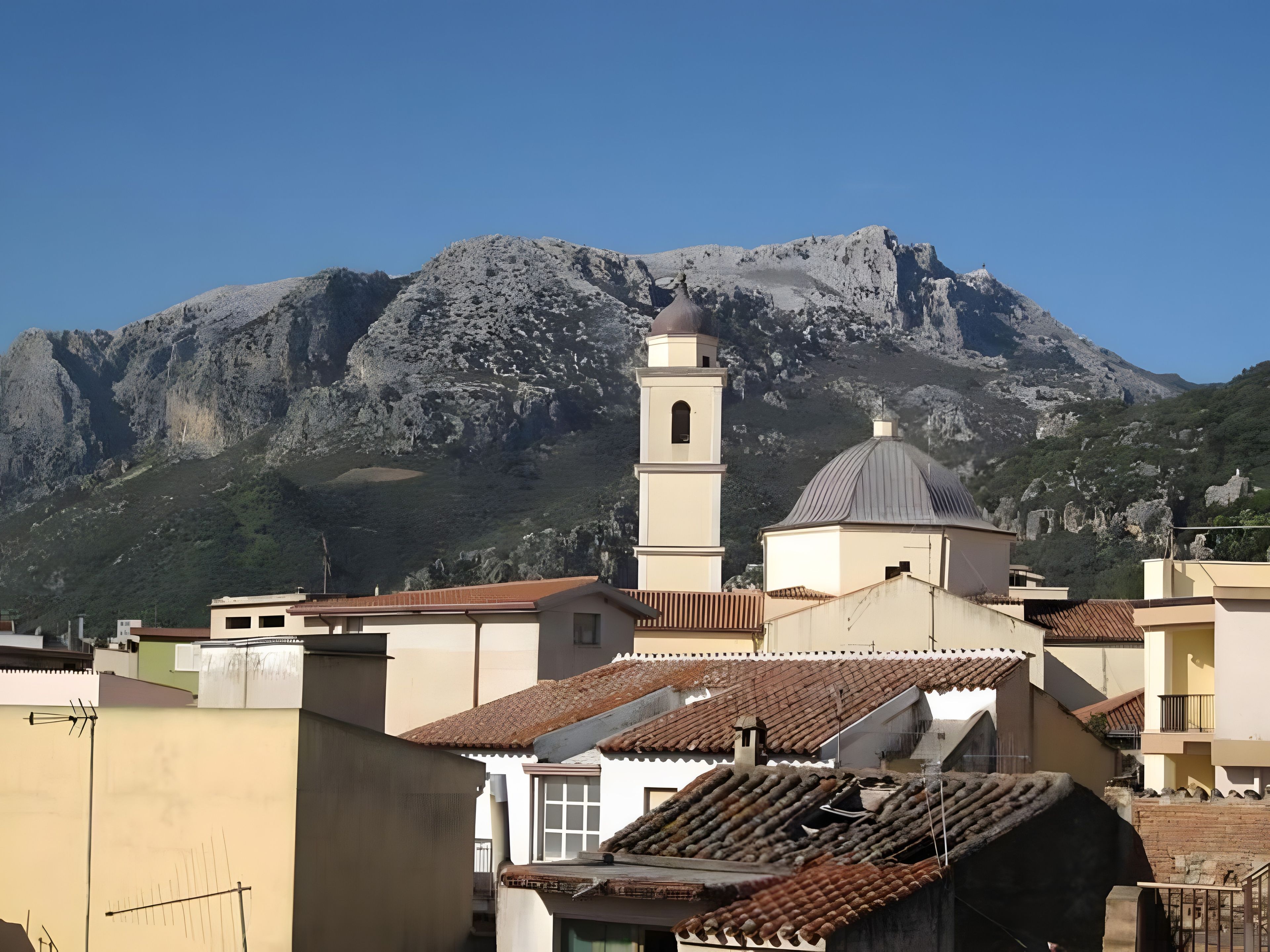 A building with a bell tower and a dome, surrounded by residential buildings and a mountain in the background.