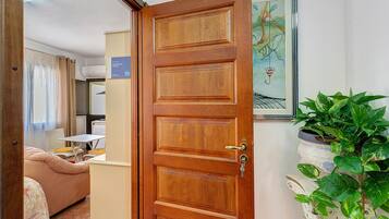 Habitación de hotel con puerta de madera, una cama, una mesita, una silla, una planta en maceta y un cuadro en la pared.